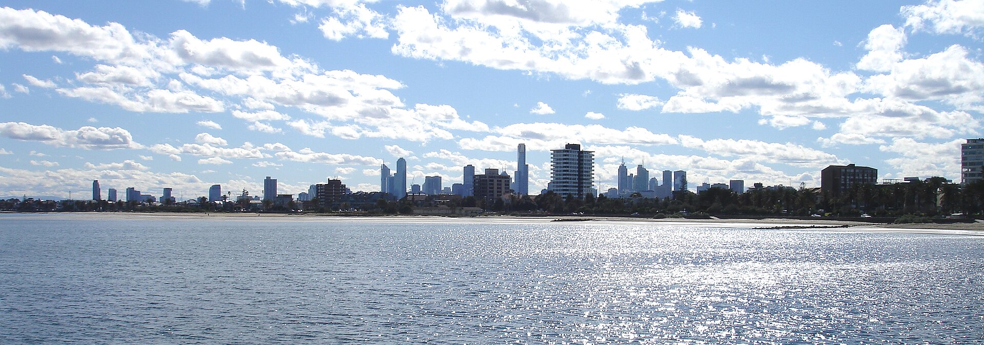 A cityscape of Melbourne rises across the water under a cloudy sky, showcasing suburban life.
